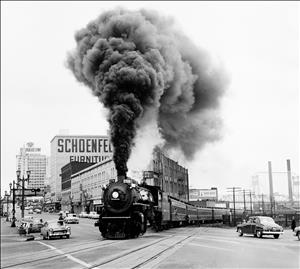 A steam locomotive rolls through a city intersection as mid-twentieth-century automobiles wait on either side. 